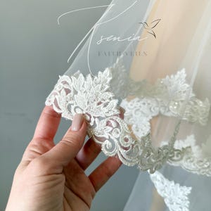 May include: Close-up of a white bridal veil with intricate floral lace trim. The delicate lace features detailed patterns and is embellished with small beads. The veil is sheer and lightweight, with the text "senia faithveils" visible.
