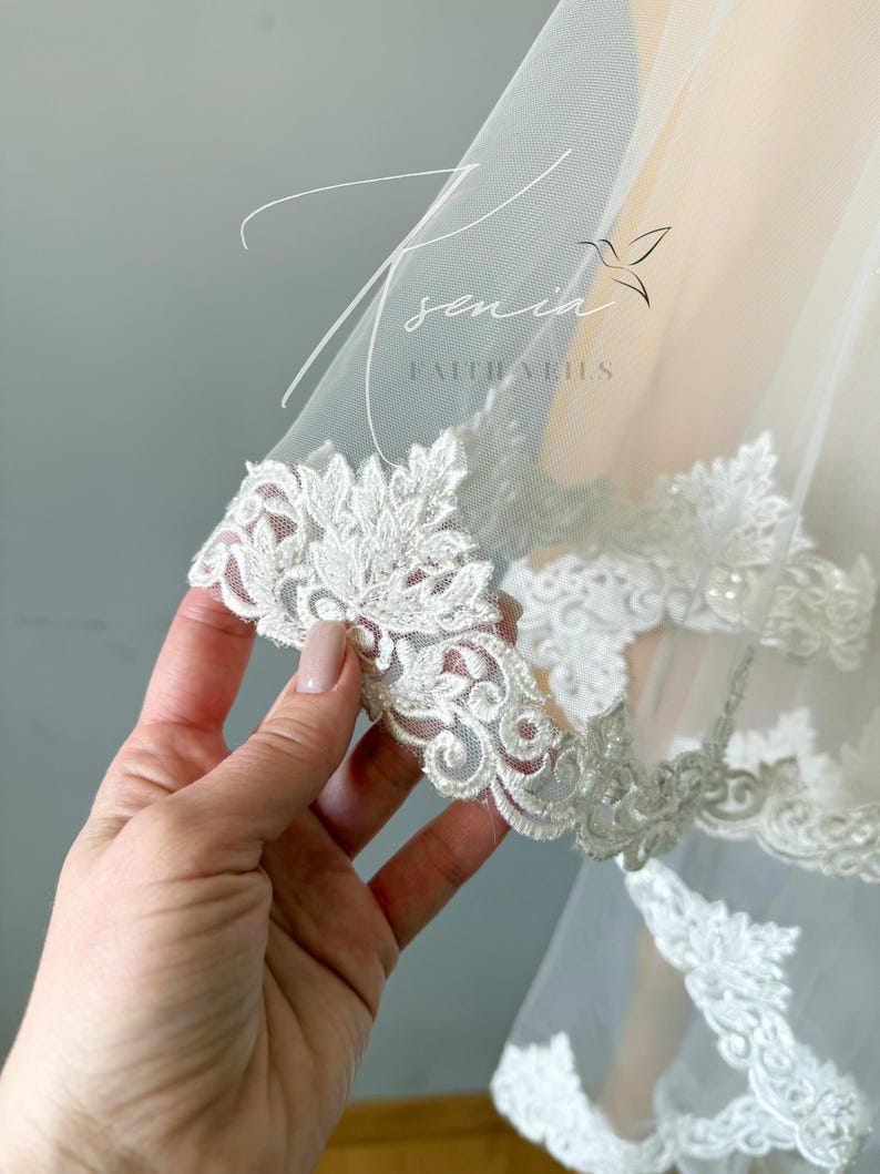 May include: Close-up of a white bridal veil with intricate floral lace trim. The delicate lace features detailed patterns and is embellished with small beads. The veil is sheer and lightweight, with the text "senia faithveils" visible.