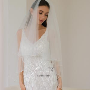 May include: A two-tiered, sheer white bridal veil with delicate beaded trim. The veil is worn over a white, embellished wedding dress. The image includes the text "senin FAITH VEILS".