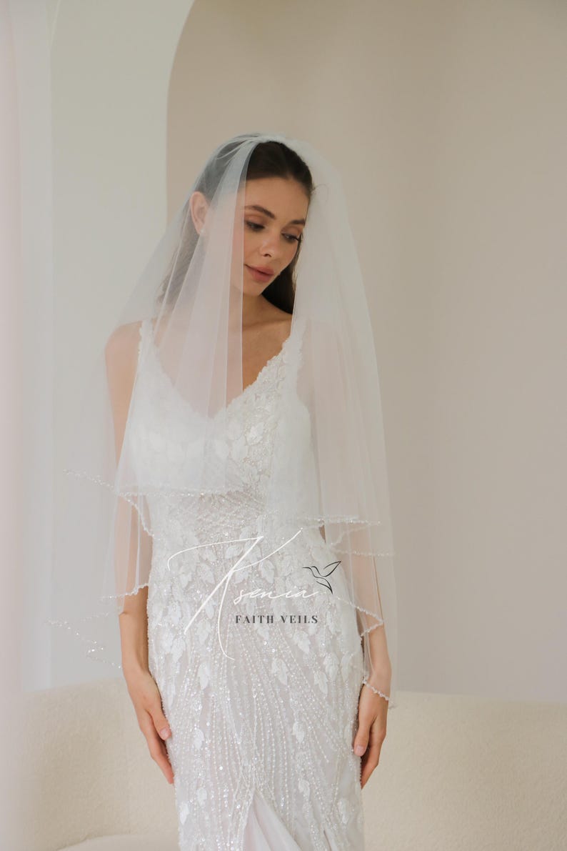 May include: A two-tiered, sheer white bridal veil with delicate beaded trim. The veil is worn over a white, embellished wedding dress. The image includes the text "senin FAITH VEILS".