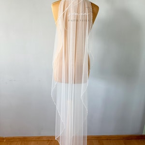 May include: A sheer white bridal veil with a delicate beaded edge and the text "senin FAITH VEILS" in a script font. The veil is displayed on a mannequin, showcasing its length and design. The veil's fabric is light and airy.