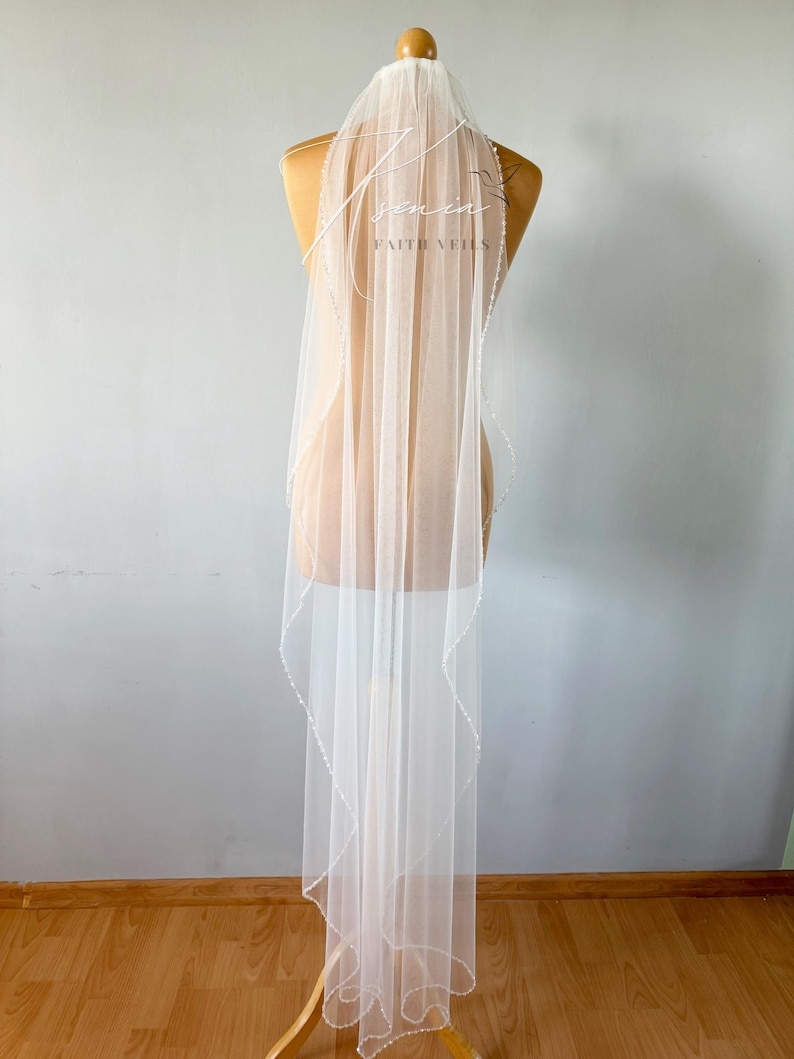 May include: A sheer white bridal veil with a delicate beaded edge and the text "senin FAITH VEILS" in a script font. The veil is displayed on a mannequin, showcasing its length and design. The veil's fabric is light and airy.