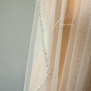 May include: A sheer white bridal veil with a delicate beaded trim. The veil is made of a soft, flowing fabric and features a strand of clear, sparkling beads along the edge. The background is a neutral color.