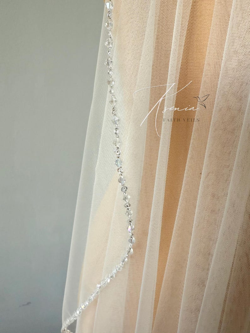 May include: A sheer white bridal veil with a delicate beaded trim. The veil is made of a soft, flowing fabric and features a strand of clear, sparkling beads along the edge. The background is a neutral color.