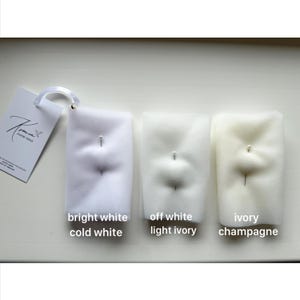May include: Three samples of bridal veil fabric in varying shades of white: bright white/cold white, off white/light ivory, and ivory/champagne. Each sample is pinned and labeled with its color name. A tag with the brand name is visible.