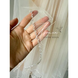 May include: A sheer white bridal veil with a delicate beaded edge. The veil is held up, showcasing the intricate details of the clear, sparkling beads. The text "Asenia Faith Veils" is visible on the veil.
