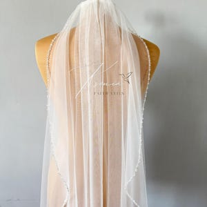 May include: A sheer white bridal veil with a beaded edge is displayed on a mannequin. The veil has the text "senia" and "FAITH VEILS" printed on it, along with a small bird graphic. The veil is made of a delicate, flowing fabric.