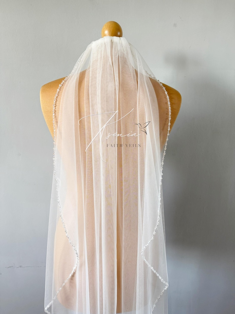 May include: A sheer white bridal veil with a beaded edge is displayed on a mannequin. The veil has the text "senia" and "FAITH VEILS" printed on it, along with a small bird graphic. The veil is made of a delicate, flowing fabric.