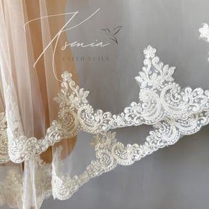 May include: Ivory-colored bridal veil with intricate, ornate lace trim. The delicate lace features floral and scrollwork patterns. The veil is sheer, allowing light to pass through, and is displayed on a mannequin. The text "Ksenia Faith Veils" is visible.
