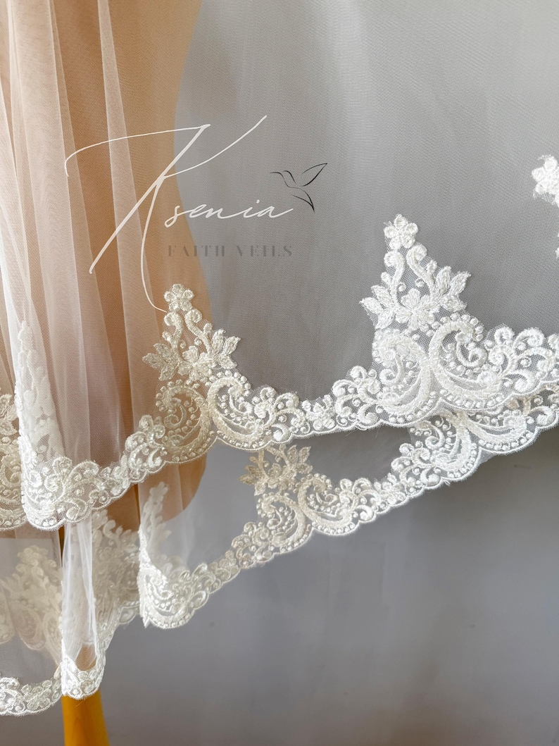 May include: Ivory-colored bridal veil with intricate, ornate lace trim. The delicate lace features floral and scrollwork patterns. The veil is sheer, allowing light to pass through, and is displayed on a mannequin. The text "Ksenia Faith Veils" is visible.