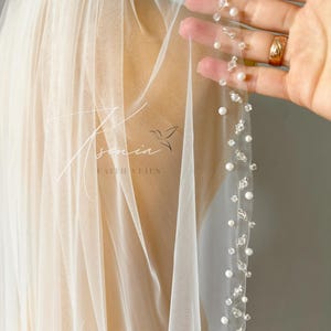 Puede incluir: Un velo de novia color marfil transparente con un borde delicado adornado con perlas blancas dispersas y cristales transparentes facetados. La tela es un tul fino y ligero. El texto Ksenia Faith Veils está impreso en el velo con una fuente blanca.