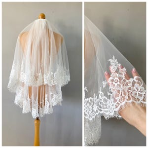 Puede incluir: Un velo de novia blanco de dos niveles hecho de tul transparente con un intrincado borde de encaje floral a lo largo de los bordes festoneados. El velo se muestra en un maniquí, destacando el diseño en capas y el bordado detallado.