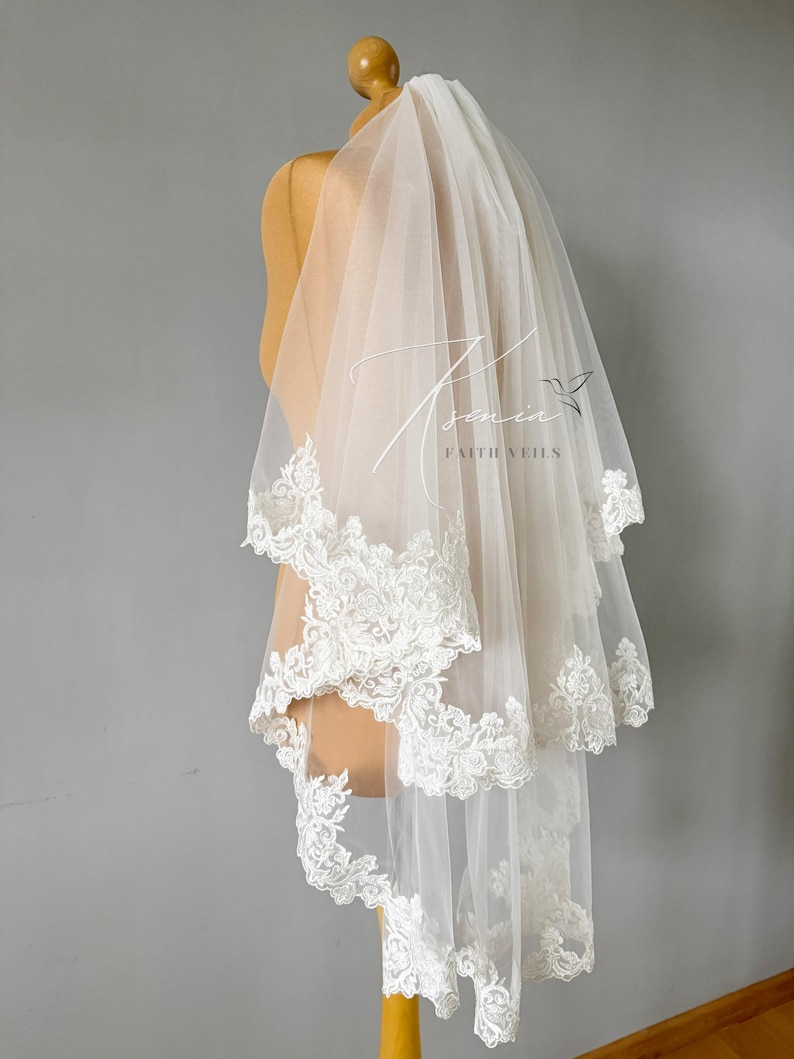 May include: A two-tiered white bridal veil with delicate lace trim. The veil is displayed on a mannequin and features the text "Tsonia Faith Veils" in a script font.
