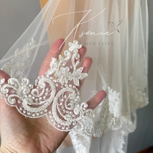 May include: A sheer white bridal veil with intricate floral lace trim. The delicate lace features detailed scrollwork and beaded accents. The veil has the text "Asenia with Veils" in a script font.