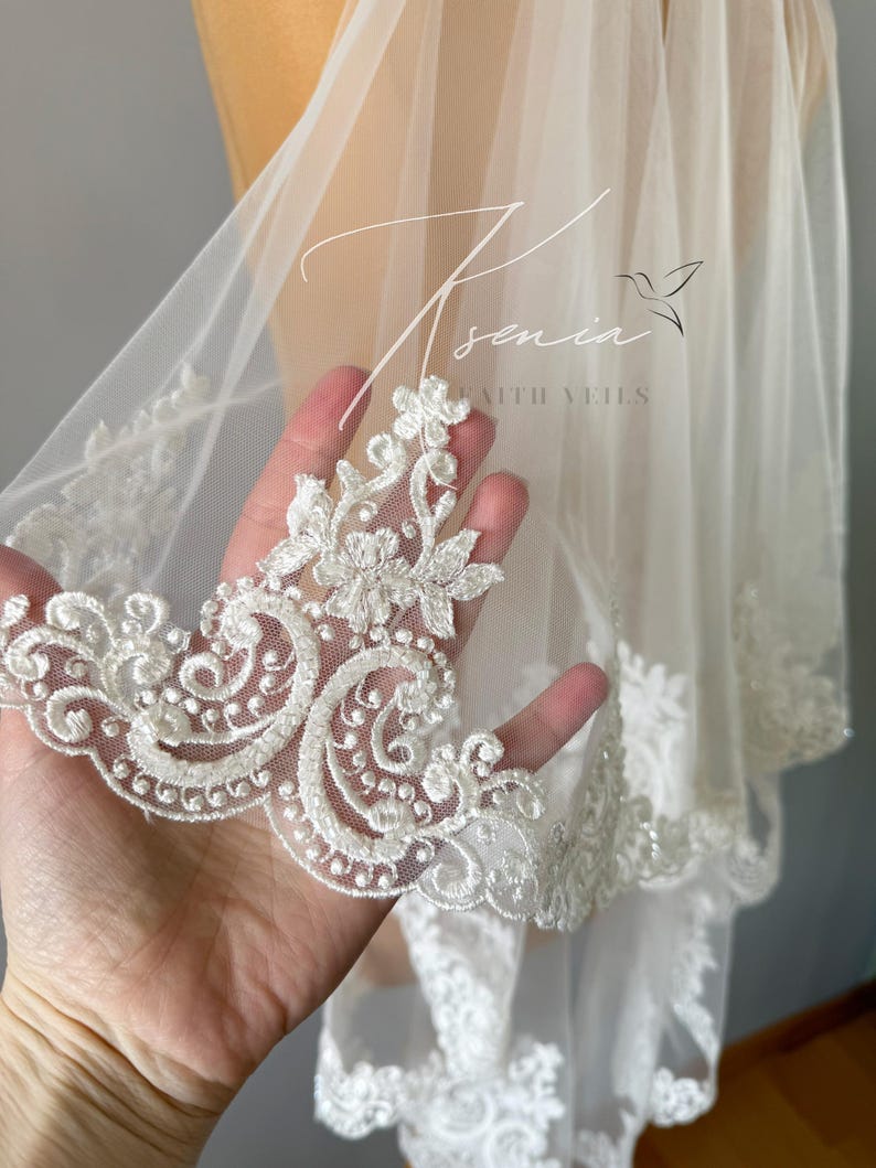 May include: A sheer white bridal veil with intricate floral lace trim. The delicate lace features detailed scrollwork and beaded accents. The veil has the text "Asenia with Veils" in a script font.