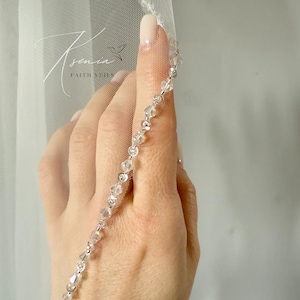 May include: A sheer white bridal veil with a delicate beaded edge. The edge is adorned with clear, sparkling crystals. The veil is held up by a hand with a light pink manicure. The text "Tsenia Faith Veils" is visible in the upper left corner.
