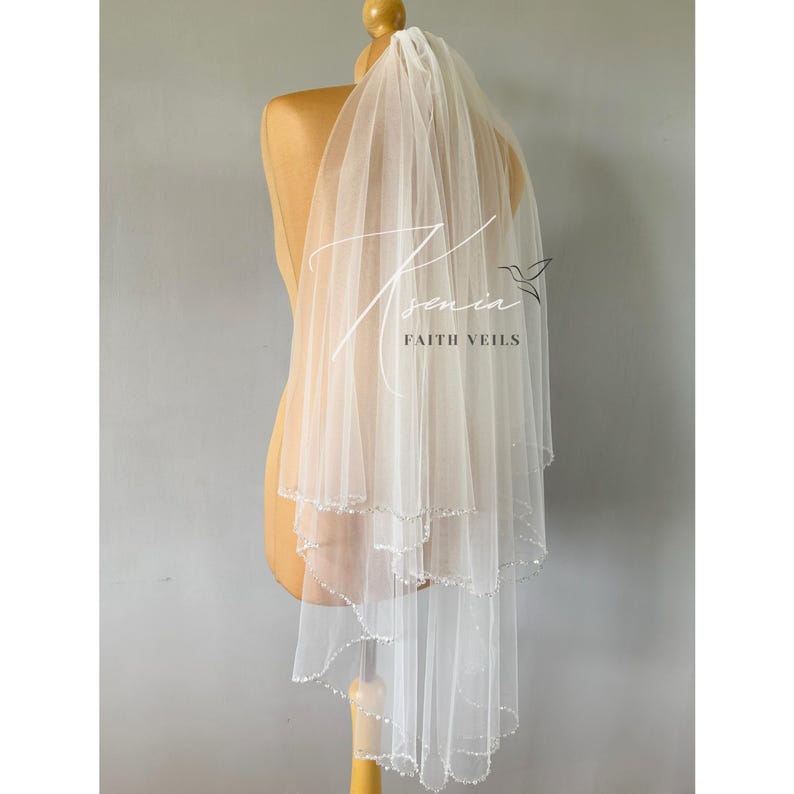 May include: A white, layered bridal veil with a beaded edge, displayed on a mannequin. The veil has a delicate, sheer appearance and features the text "Asenia Faith Veils" in a script font.