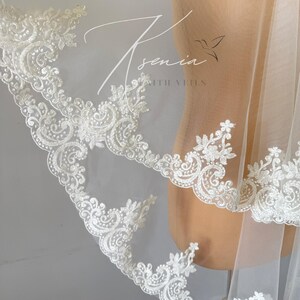 May include: A white bridal veil with intricate floral lace detailing along the edges. The delicate lace features swirling patterns and small, raised embellishments. The veil is sheer, allowing light to pass through, and is draped over a mannequin.