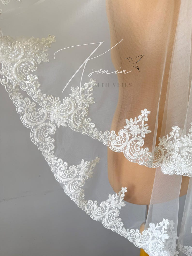 May include: A white bridal veil with intricate floral lace detailing along the edges. The delicate lace features swirling patterns and small, raised embellishments. The veil is sheer, allowing light to pass through, and is draped over a mannequin.