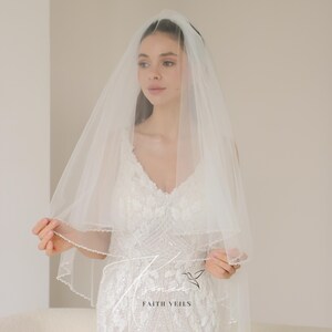 May include: A white bridal veil with a beaded edge, worn over a white wedding dress. The veil is sheer and layered, with the text "FAITH VEILS" visible. The dress has a V-neck and intricate floral embellishments.