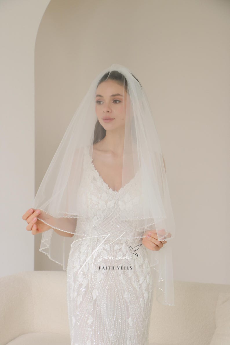 May include: A white bridal veil with a beaded edge, worn over a white wedding dress. The veil is sheer and layered, with the text "FAITH VEILS" visible. The dress has a V-neck and intricate floral embellishments.
