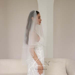 May include: A bride wearing a white wedding veil with a beaded edge. The veil is layered and sheer, complementing the white, embellished wedding dress. The image is taken indoors with a neutral background. The text "Faith Veils" is visible.