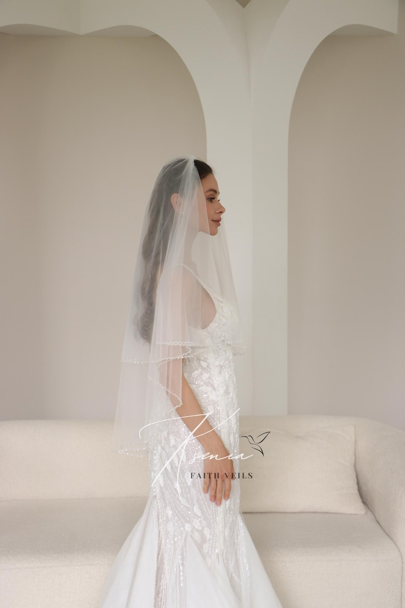 May include: A bride wearing a white wedding veil with a beaded edge. The veil is layered and sheer, complementing the white, embellished wedding dress. The image is taken indoors with a neutral background. The text "Faith Veils" is visible.