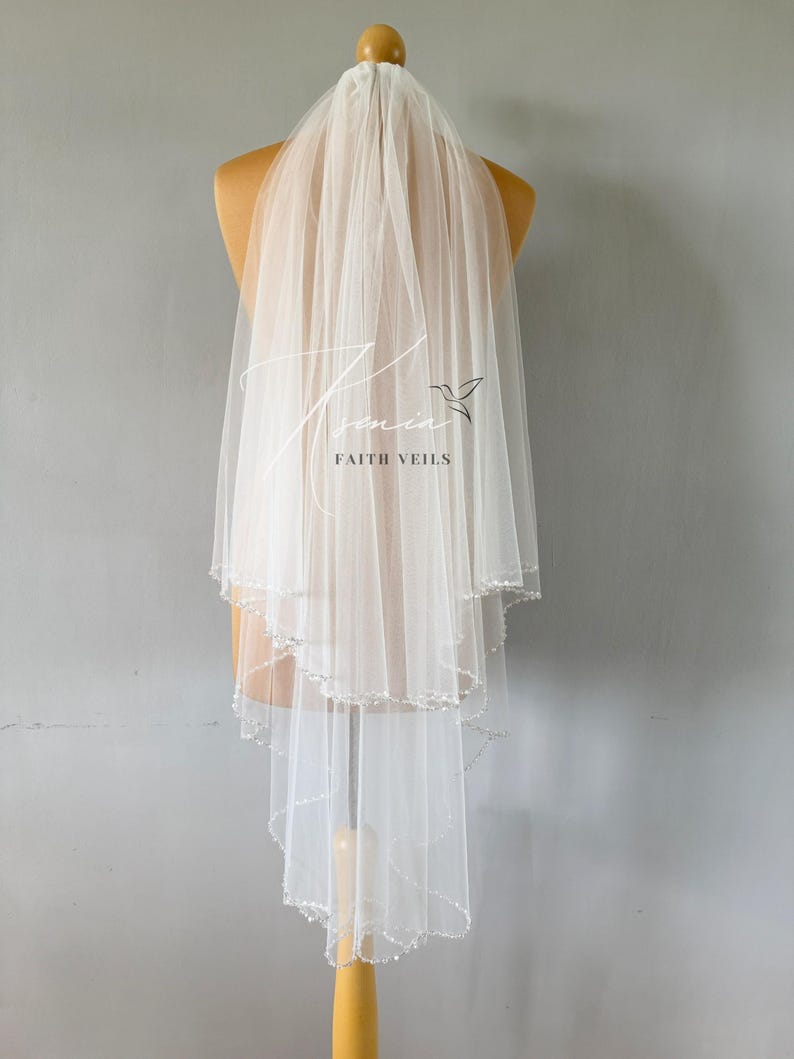 May include: A white, layered bridal veil with a scalloped edge and beaded trim. The veil is displayed on a mannequin and features the text "Tamin Faith Veils". The veil is made of a sheer, lightweight fabric.