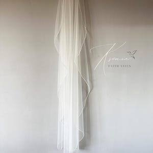 May include: A sheer white bridal veil with a delicate beaded edge hangs against a gray wall. The veil is long and flowing, with a soft, ethereal appearance. The text "Ksenia Faith Veils" is visible in the image.