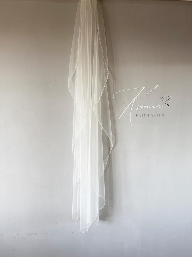 May include: A sheer white bridal veil with a delicate beaded edge hangs against a gray wall. The veil is long and flowing, with a soft, ethereal appearance. The text "Ksenia Faith Veils" is visible in the image.