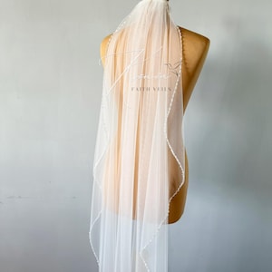 May include: A sheer white bridal veil with a delicate beaded edge and the text "Faith Veils" in elegant script. The veil is draped over a mannequin, showcasing its flowing design and intricate details. The veil's length is approximately 72 inches.