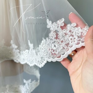 May include: A white bridal veil with a delicate floral lace trim. The veil is made of sheer fabric and features the text "Xenia Faith Veils" in a script font. The lace trim is adorned with small, detailed flower designs.