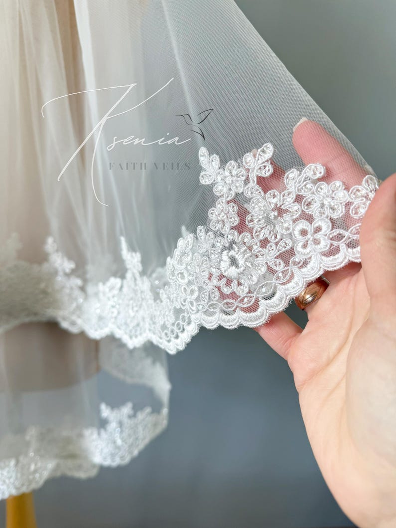 May include: A white bridal veil with a delicate floral lace trim. The veil is made of sheer fabric and features the text "Xenia Faith Veils" in a script font. The lace trim is adorned with small, detailed flower designs.
