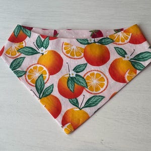 Może przedstawiać: R&oacute;żowa bandana z powtarzającym się wzorem pomarańczy, plasterk&oacute;w pomarańczy i zielonych liści. Pomarańcze są w żywym pomarańczowym kolorze z czerwonymi akcentami, a liście są w głębokiej zieleni. Bandana jest wykonana z miękkiej tkaniny.