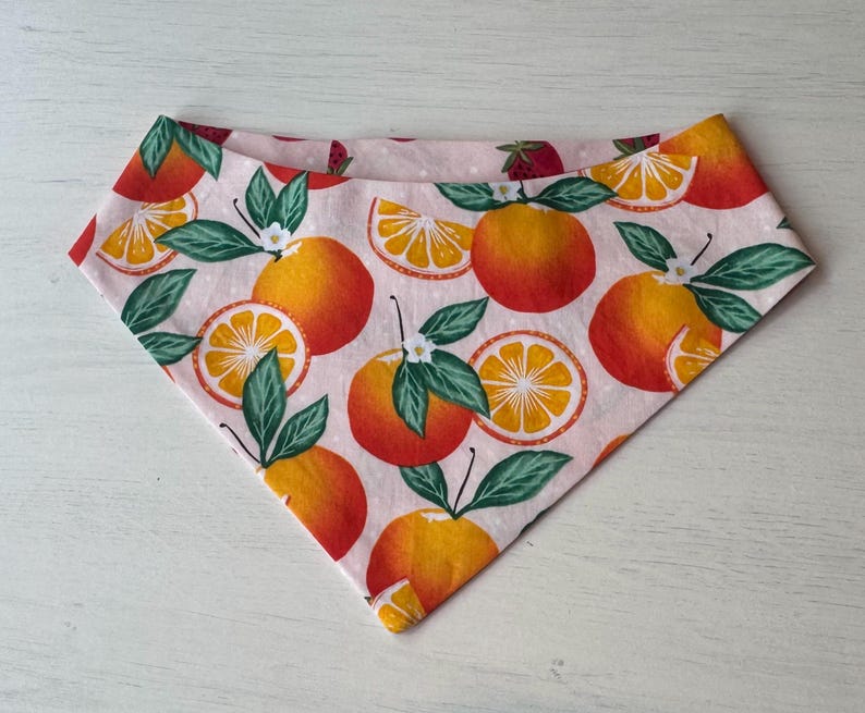 Może przedstawiać: R&oacute;żowa bandana z powtarzającym się wzorem pomarańczy, plasterk&oacute;w pomarańczy i zielonych liści. Pomarańcze są w żywym pomarańczowym kolorze z czerwonymi akcentami, a liście są w głębokiej zieleni. Bandana jest wykonana z miękkiej tkaniny.