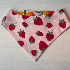 Może przedstawiać: R&oacute;żowa bandana dla psa z wzorem truskawek. Bandana ma duże czerwone truskawki z zielonymi łodygami i liśćmi na jasnor&oacute;żowym tle. Druga strona ma wz&oacute;r ż&oacute;łtych i pomarańczowych owoc&oacute;w.