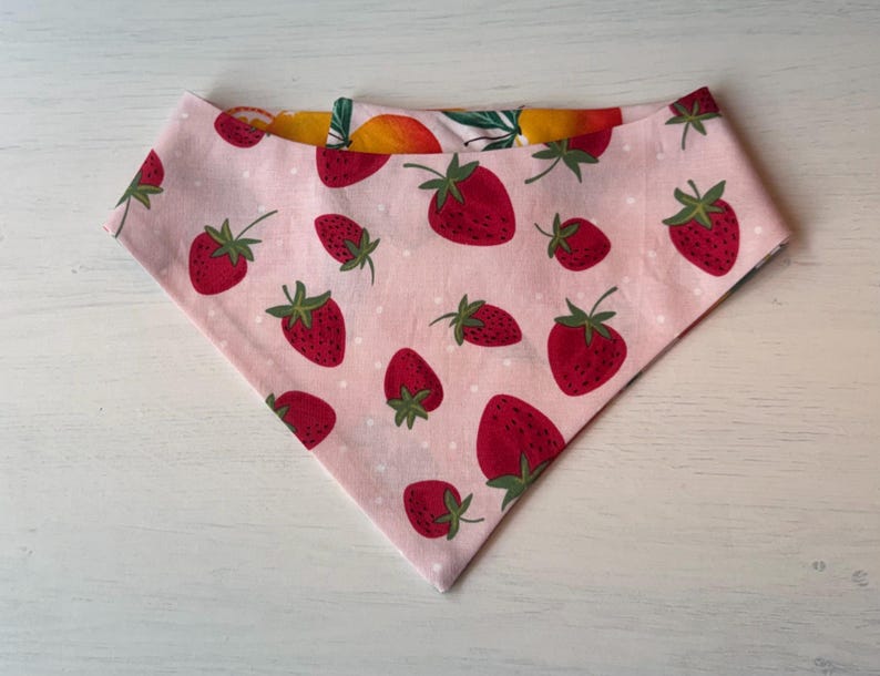 Może przedstawiać: R&oacute;żowa bandana dla psa z wzorem truskawek. Bandana ma duże czerwone truskawki z zielonymi łodygami i liśćmi na jasnor&oacute;żowym tle. Druga strona ma wz&oacute;r ż&oacute;łtych i pomarańczowych owoc&oacute;w.