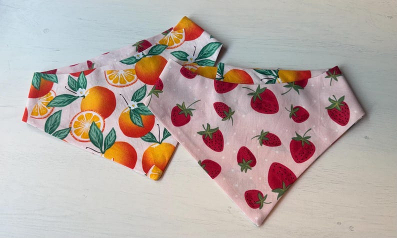 Może przedstawiać: Dwie tr&oacute;jkątne bandany z owocowymi wzorami. Jedna przedstawia pomarańcze z zielonymi liśćmi na białym tle. Druga ma czerwone truskawki z zielonymi łodygami na r&oacute;żowym tle w kropki. Obie są wykonane z tkaniny.