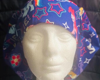 May include: Blue surgical scrub hat with a colorful My Little Pony print featuring stars, rainbows, and ponies. The hat has a tie at the back.