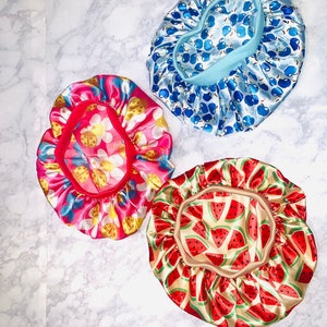 May include: Three satin hair bonnets with different patterns. One bonnet is blue with a white and blue apple pattern, one is pink with a white and yellow flower and ladybug pattern, and one is beige with a red and green watermelon pattern.