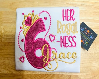 Peut inclure: Un t-shirt blanc avec des broderies roses et dorées. La broderie représente un grand chiffre six rose avec des paillettes, une couronne dorée et le texte "HER ROYAL-NESS Grace".