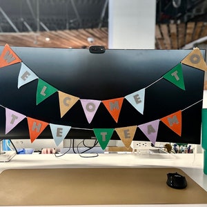 May include: A colorful felt banner with the words "Welcome to the Team" hanging on a black computer monitor. The banner is strung with white thread and has a green, orange, light blue, purple, and brown color scheme.