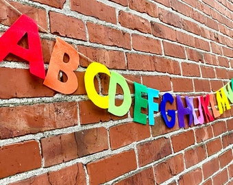 Könnte beinhalten: Ein farbenfroher Filzbanner mit den Buchstaben des Alphabets, beginnend mit A und endend mit Z. Die Buchstaben sind mit einer Schnur aneinandergereiht und hängen an einer Backsteinwand.