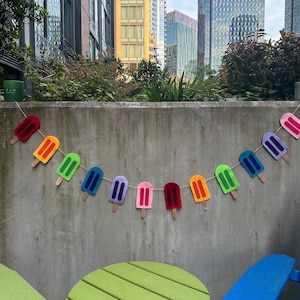 May include: A vibrant popsicle garland featuring felt ice pops in a rainbow of colours: red, orange, green, blue, purple, and pink. Each ice pop has a wooden stick and is strung on twine. The garland is displayed against a concrete wall.