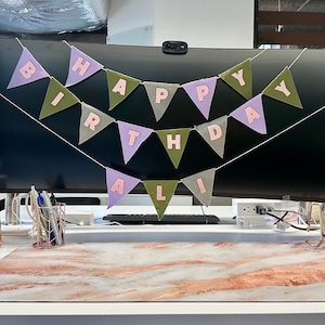 May include: A pink and green felt banner with the words "Happy Birthday Ali" hanging in front of a black computer monitor.