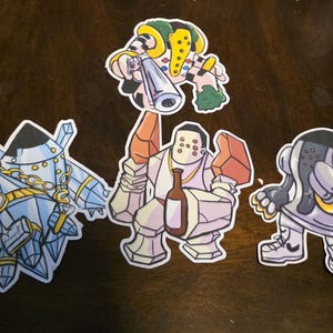 May include: Three die-cut vinyl stickers featuring stylized robotic characters. The left sticker shows a crystal-like figure with a gold chain. The center sticker depicts a stone-like figure holding a bottle. The right sticker shows a rounded figure.