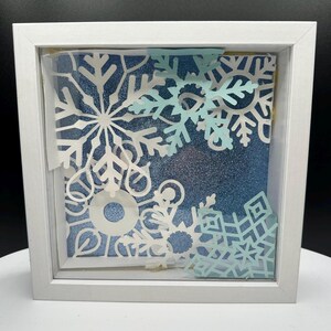 May include: A square white shadow box frame containing layered paper cutouts of various snowflake shapes in white and light blue. The background is a textured, glittery blue surface, creating a multi-dimensional effect within the frame.