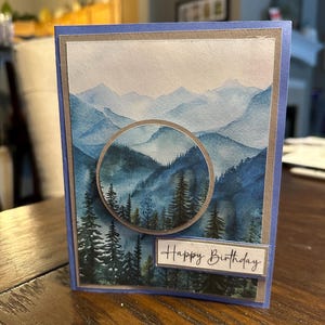 May include: A handmade birthday card featuring a watercolor landscape of blue mountains and evergreen trees. The card has a blue border and a circular cutout showcasing the mountain scene. The words "Happy Birthday" are printed on a rectangular label.