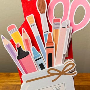Peut inclure: Une découpe en papier en forme de maison rouge remplie d'illustrations de fournitures artistiques, dont un pinceau, des ciseaux, des crayons, un marqueur et un crayon de cire. Un bocal blanc affiche le calendrier de janvier 2027.