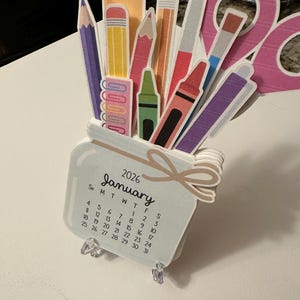 May include: A papercraft calendar for January 2026, shaped like a jar filled with school supplies. The jar contains pencils, crayons, a ruler, and a notepad. The calendar is supported by clear plastic stands.